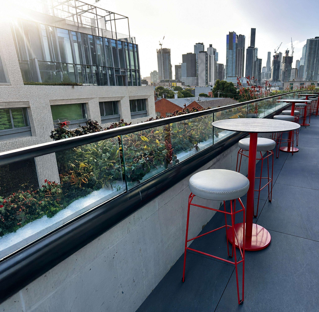 A rooftop terrace with modern red bar stools and round white-top tables, overlooking a city skyline with glass buildings and greenery along the railing.