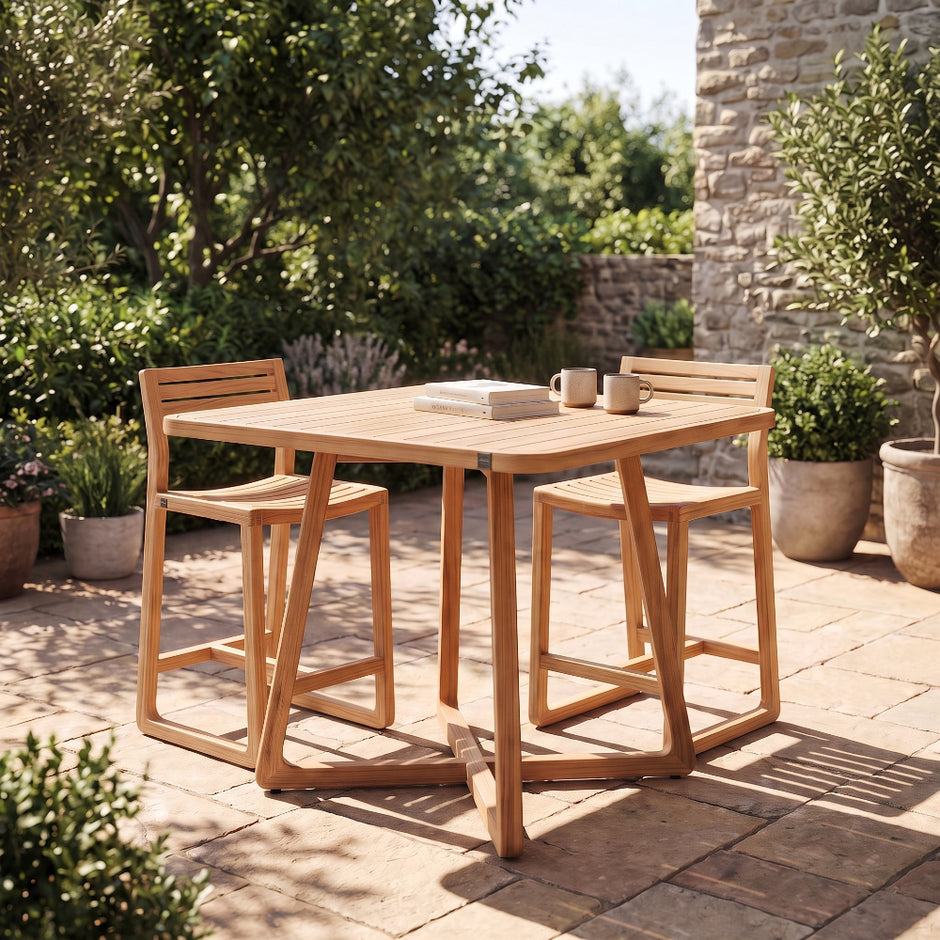 A square outdoor dining set with a light natural wood table and four matching bar-height stools, placed on a stone patio in a garden setting.