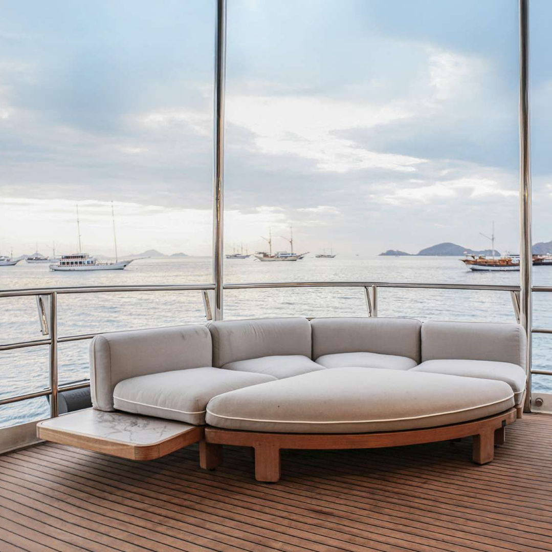 A curved outdoor lounge sofa with light beige cushions and a wooden base sits on a yacht deck with large windows overlooking a calm sea and boats.